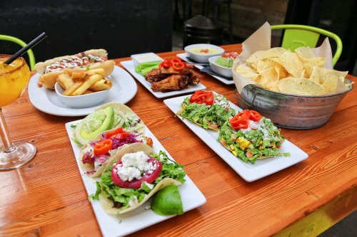 A vibrant spread of tacos, wings, chips, and a drink on a wooden table, showcasing colorful and fresh ingredients.