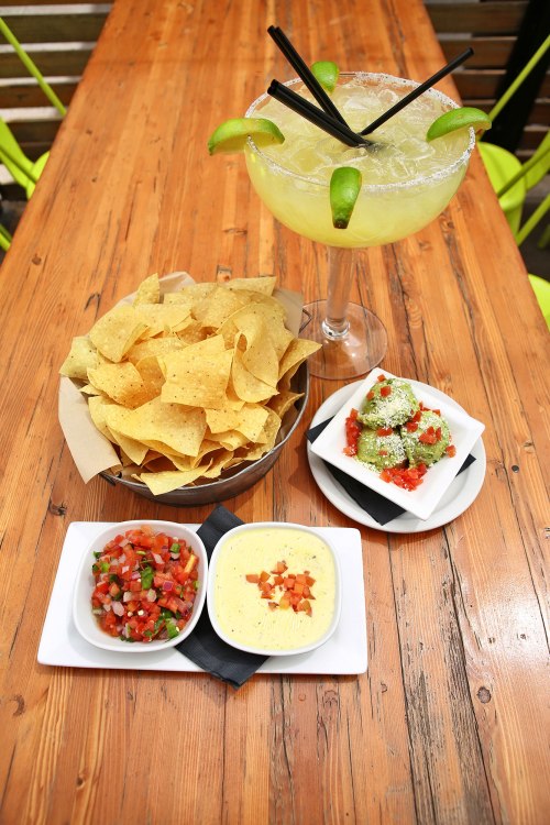 A wooden table with a large margarita, tortilla chips, salsa, and cheese dip arranged neatly.