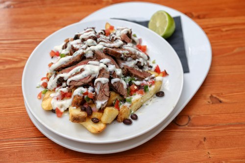 A plate of loaded fries topped with grilled steak, diced tomatoes, black beans, and drizzled with sauce, served with a lime wedge.