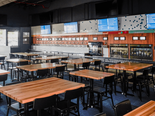 A spacious bar area with wooden tables and a wall of taps, featuring a modern design and multiple screens.