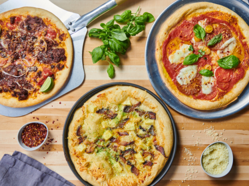 Three pizzas on a wooden table: one with meat, one with tomatoes and cheese, and one with pineapple and ham, garnished with basil.