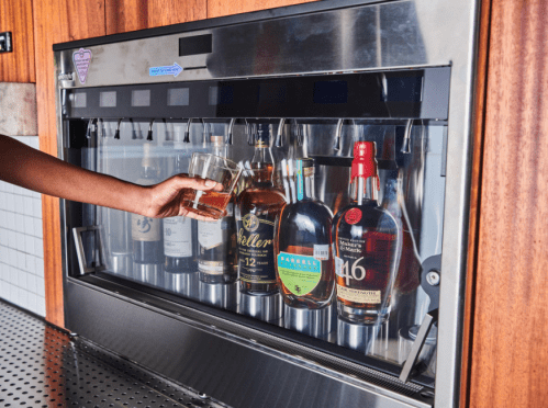 A hand reaches for a glass from a modern liquor dispenser showcasing various whiskey bottles.