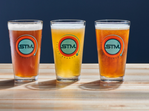 Three glasses of beer in varying colors, each with a circular STM logo, placed on a wooden table against a blue background.