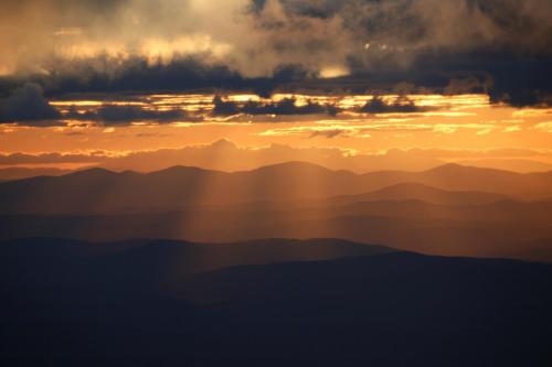 Sunset over layered mountains, with dramatic clouds and rays of light illuminating the landscape.