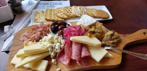 A wooden platter with assorted cheeses, meats, olives, pickled vegetables, mustard, and a side of crackers.