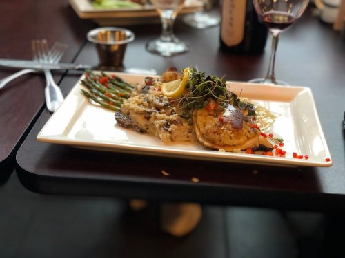A plated dish featuring fish, asparagus, and garnishes, served on a white rectangular plate with wine glasses in the background.