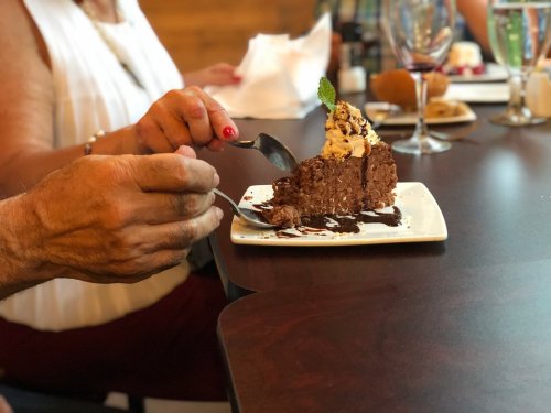 A hand holding a fork and knife, preparing to cut a slice of chocolate cake topped with cream and a mint leaf.