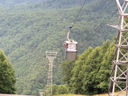A cable car travels over a lush green mountain landscape, with power lines visible in the foreground.