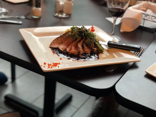 A plated dish of sliced meat garnished with greens, served with a dark sauce, on a table with a glass of red wine.