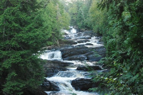 A serene river flows over rocky terrain, surrounded by lush green trees and gentle cascades of water.