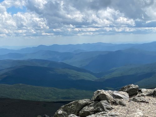 A panoramic view of rolling mountains under a cloudy sky, with rocky foreground and lush green valleys below.