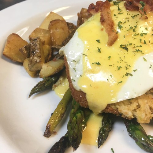 A plate featuring a breaded dish topped with a poached egg and hollandaise sauce, served with asparagus and roasted potatoes.