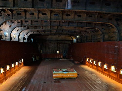 Interior of a large, empty ship hold with rusted metal beams and dim lighting along the walls.