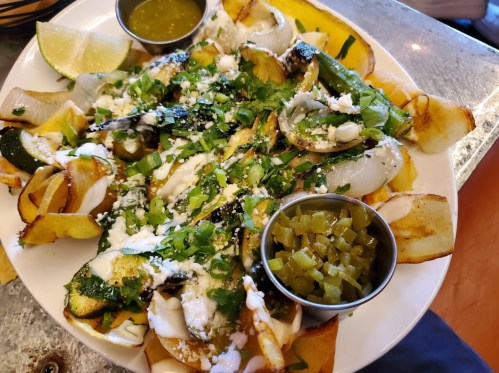 A plate of grilled vegetables topped with cheese, cilantro, and sauces, served with lime wedges and pickled jalapeños.