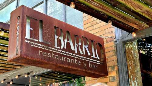 Sign for "El Barrio" restaurant and bar, featuring a rustic metal design, hanging above a brick facade.