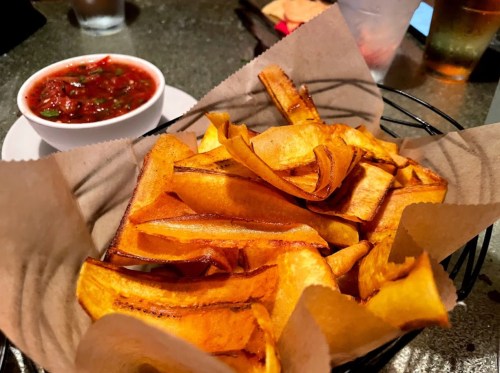 A basket of crispy plantain chips served with a small bowl of salsa on a table.