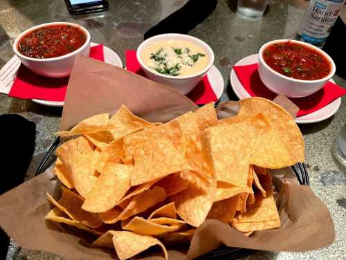 A basket of tortilla chips with two bowls of salsa and one bowl of creamy dip on a table.