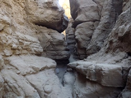 Narrow canyon with steep, rocky walls and sandy ground, illuminated by sunlight from above.