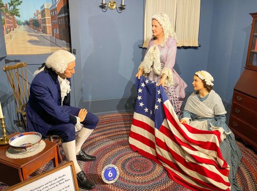 A historical scene with three figures in period clothing, one holding an American flag, set in a room with a painted backdrop.