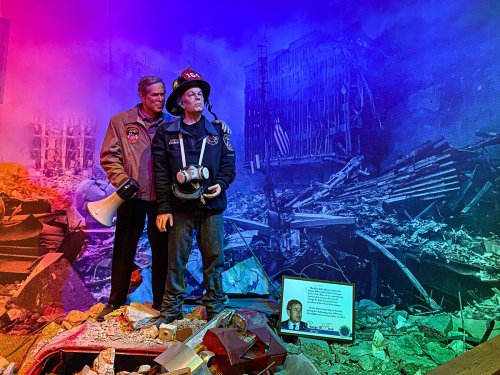 A firefighter stands among rubble, with a backdrop of a destroyed building and a tribute plaque nearby.