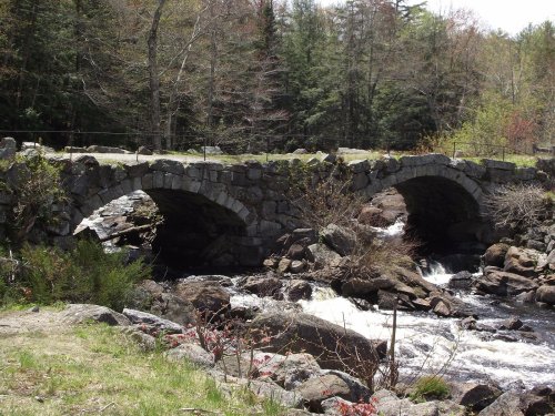 A stone bridge with two arches spans a rushing river, surrounded by trees and rocky terrain.