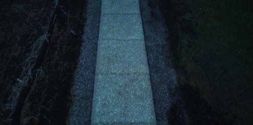A dimly lit pathway made of concrete slabs, bordered by gravel and dark soil, creating a serene nighttime scene.