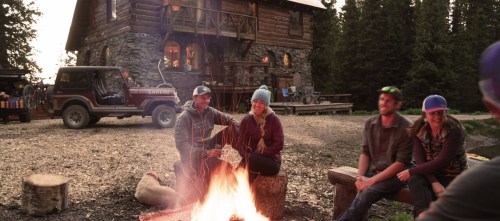 A group of four people sits around a campfire in front of a rustic cabin, enjoying a cozy evening in nature.