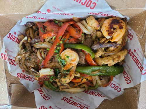 A takeout box filled with grilled shrimp, beef, bell peppers, onions, and herbs on a bed of seasoned vegetables.