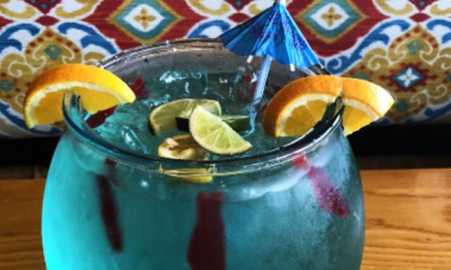 A colorful cocktail in a fishbowl glass, garnished with orange and lime slices, and a small blue umbrella.