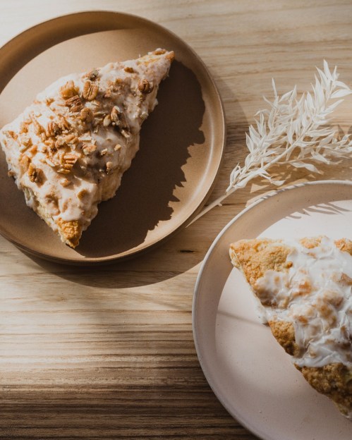 Two scones on minimalist plates, one topped with icing and nuts, accompanied by a dried plant on a wooden surface.