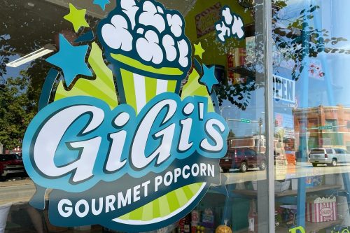 Sign for GiGi's Gourmet Popcorn featuring a colorful popcorn graphic, visible through a shop window.