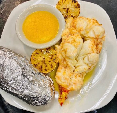 A plate featuring grilled lobster tail, charred lemon halves, and a baked potato wrapped in foil, with a dipping sauce.