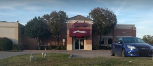 A restaurant building with a maroon awning and a blue car parked in front, surrounded by trees and grass.