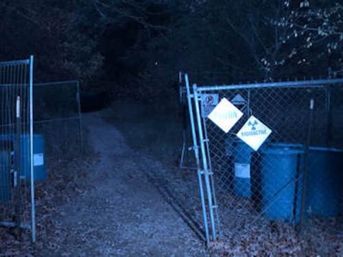 A dimly lit path leads through a chain-link fence with warning signs and blue barrels, surrounded by trees.