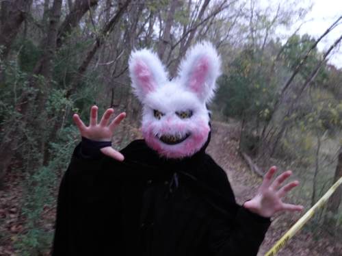 A figure in a creepy bunny mask and black cloak poses menacingly in a wooded area.