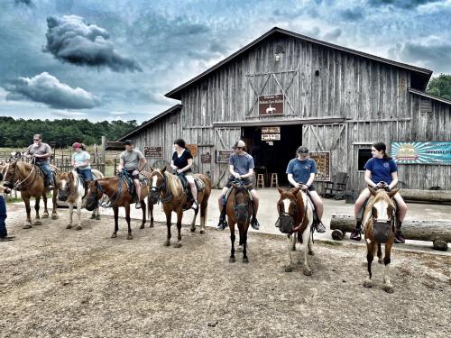 A group of people on horseback in front of a rustic barn, with a cloudy sky overhead.