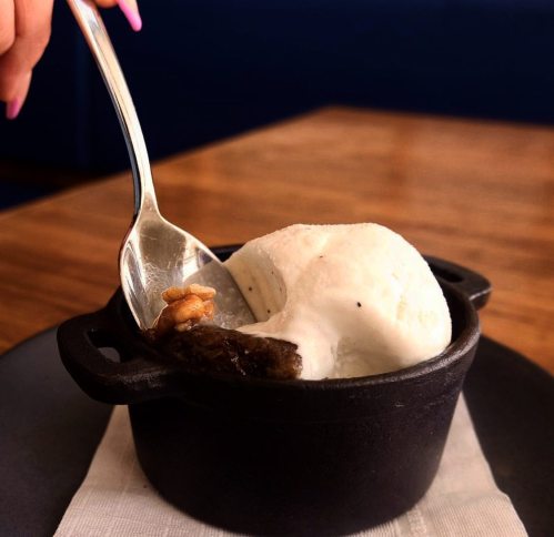 A spoon scoops vanilla ice cream from a small black pot, with a chocolate dessert underneath and a walnut on top.