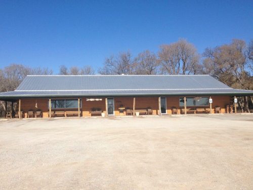 A single-story wooden building with a metal roof, surrounded by trees and gravel, under a clear blue sky.