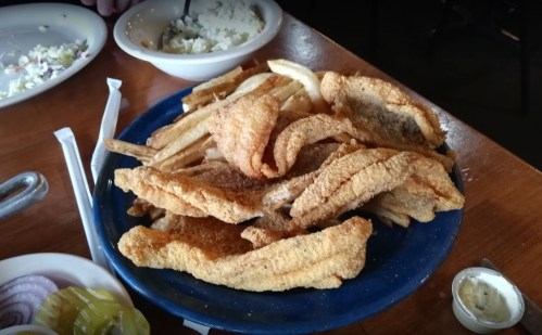 A blue plate piled high with golden-brown fried fish and crispy fries, with side dishes in the background.