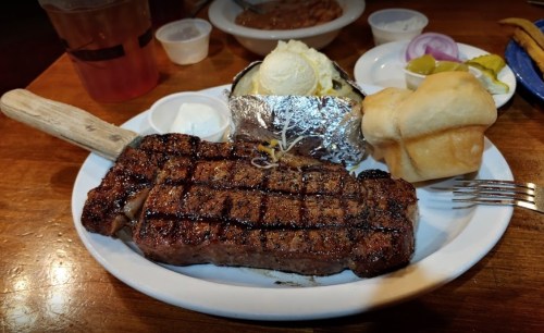 A plate featuring a grilled steak, baked potato with toppings, a roll, and condiments on a wooden table.
