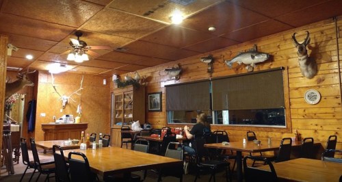 A rustic restaurant interior with wooden walls, mounted fish and deer heads, and empty tables. A person sits at a table.