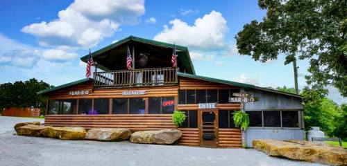 A rustic barbecue restaurant with a wooden exterior, flags, and a welcoming "Open" sign under a blue sky.