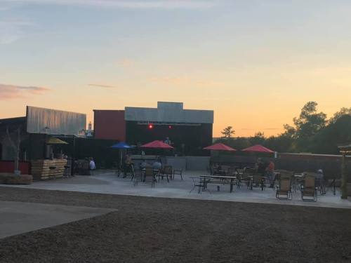 Outdoor venue at sunset with tables, chairs, and a stage in the background, surrounded by trees.