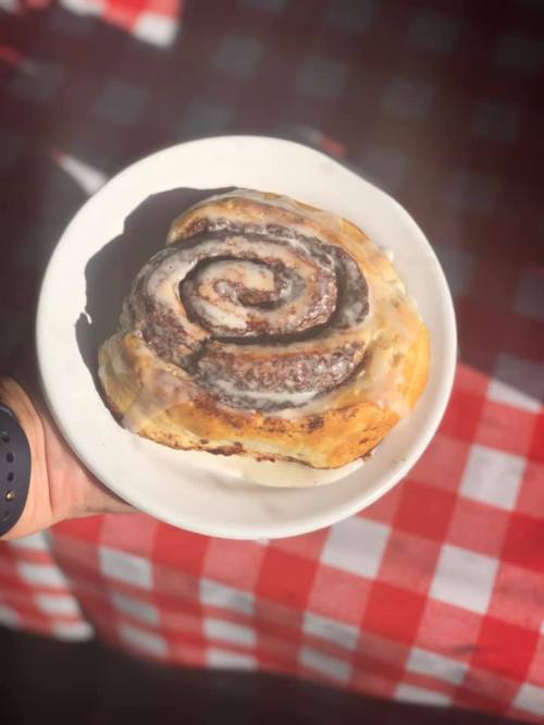A hand holding a cinnamon roll on a white plate, with a checkered red and white tablecloth in the background.