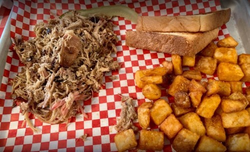 A plate with pulled pork, golden-brown diced potatoes, and two slices of toasted bread on a red and white checkered paper.