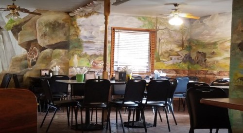 A cozy restaurant interior featuring a mural of nature, with empty tables and chairs arranged for dining.