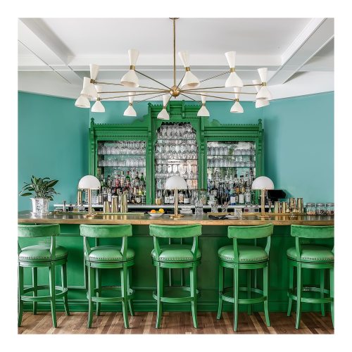A stylish green bar with high stools, a decorative back bar, and modern lighting against a teal wall.