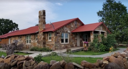 A rustic stone house with a red metal roof, surrounded by greenery and a stone pathway.