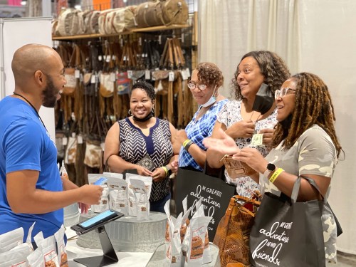 A group of four women engage with a vendor at a market, smiling and holding bags while discussing products.