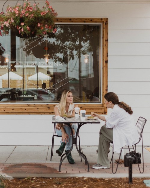 Two women sit at a small outdoor table, enjoying coffee and food, with a café window and hanging flowers in the background.